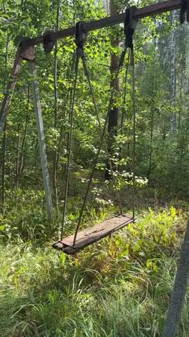 The video shows a rusty metal swing set in a wooded area. The swing seat is made of wood and appears to be old and worn. The swing is empty and not in motion. The surrounding environment is overgrown with grass and trees, suggesting that the area may be abandoned or neglected. The lighting suggests it is daytime, with sunlight filtering through the trees.