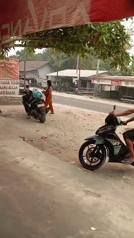 Two individuals, appearing to be two males, one older and one younger, are seated on a dark-colored motorcycle. The older individual is driving, and the younger is seated behind him. They are moving away from the viewer on a dirt and gravel road. To the left, a woman in an orange dress stands next to a parked dark-colored motorcycle. Behind them, buildings and trees are visible.
