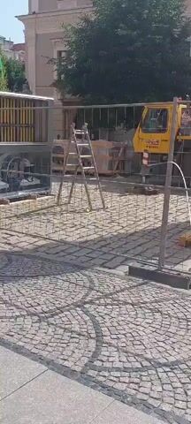 A construction site is visible through a chain-link fence. A yellow crane truck with a red boom is parked in the center of the frame. Several large, wrapped items are on wooden pallets in the foreground. A ladder is leaning against some construction materials on the left side of the frame. The ground is paved with cobblestones. A building with a light-colored facade and windows is visible in the background. A large tree is situated in front of the building. The sky is clear and blue.