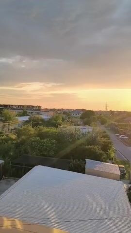 This is a vertical video of a sunset over a suburban landscape. The sun is a bright, glowing orb in the sky, casting a warm orange and yellow light over the scene. The sky is partly cloudy, with some darker clouds to the left and lighter, more wispy clouds to the right. Below the sky, there are rooftops of houses, trees, and a road. A white metal roof of a building is visible in the foreground. The overall impression is one of a peaceful and beautiful evening.