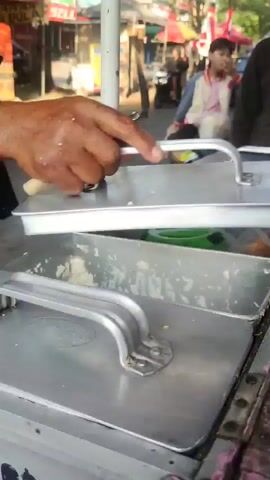 A person's hands are seen preparing food from a stainless steel cart. The person is holding a red and white bowl and uses a wooden utensil to scoop something from a bin on the cart into the bowl. The background appears to be a busy outdoor market with people and vehicles. The person speaks and receives payment.