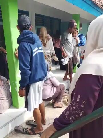 The video depicts a scene outside a building, likely a school or community center, in what appears to be a warm, sunny day. Several people are gathered on a tiled patio area. Most of the women are wearing hijabs. Some of the individuals are sitting on the edge of the patio, while others are standing around talking. A young man wearing a navy blue hoodie with white lettering on the back, gray shorts, and a baseball cap, is standing with his back to the camera. Several pairs of shoes and sandals are scattered on the patio. There is a rack of shoes near the entrance of the building. The building has green support pillars and blue window frames. A man carries a sleeping child, while another child is near him. Overall, the scene seems to show a gathering of people, possibly waiting for something or after an event.