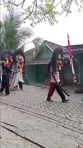Three figures in elaborate costumes resembling mythical creatures, possibly Barongan from Indonesian folklore, are walking down a paved pathway. The figures have large, exaggerated masks with fierce expressions, long black hair, and bodies covered in fabric. One figure has a distinctive black and white striped pattern on its lower body. A younger individual, dressed in black pants and a white shirt with a red and green sash, walks ahead of two of the costumed figures, holding a decorative item that resembles a spear or wand. Another person, dressed in casual clothing, walks to the left of the costumed figures. In the background, there is a building with a green wall and a red tiled roof, and trees with green leaves overhead. A motorcycle is parked in front of the building. An Indonesian flag, red and white, is visible on a pole behind one of the costumed figures. The ground is paved with bricks, and there is a ditch or cracked area in the foreground. The sky is overcast. The figures appear to be participating in a procession or performance.