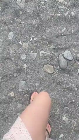 The video shows a first-person perspective of someone walking on a rocky, sandy path. The path is gray and scattered with small stones and some dried vegetation. The person is wearing a light pink, lace-like cover-up over a bathing suit and flip-flops. The camera is pointed downwards, focusing on the path and the person's feet as they walk. The overall impression is of a casual walk along a beach or a similar natural environment.