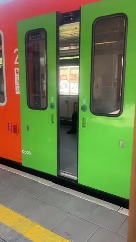 A train with orange and green carriages is stopped at a station platform. The doors of one carriage are open, revealing the interior. On the orange section of the carriage, the number '2' is displayed along with symbols indicating accessibility for passengers with children, those with disabilities, and cyclists. A yellow and black striped safety line runs along the edge of the platform. The train appears to be in motion, with the camera tracking alongside it.