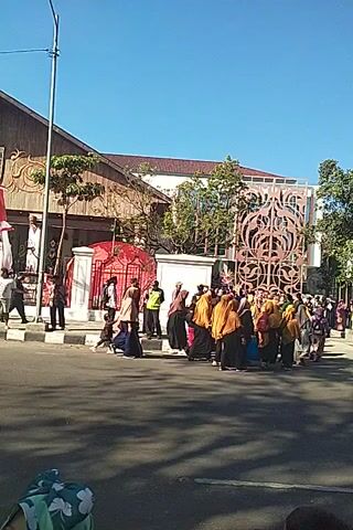 A crowd of people dressed in yellow and brown uniforms are standing in a line. Some of the people are wearing headscarves. There is a building in the background with an ornate design on it. The sky is blue and clear.