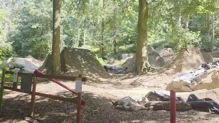 The video shows a dirt bike park in a wooded area. There are several dirt jumps of various sizes and shapes, some covered with tarps. The ground is covered in dirt, gravel, and some rocks. There are trees and foliage surrounding the park, and sunlight filters through the leaves. In the foreground, there is a red fence and some red poles. There are no people or animals visible in the video. The video appears to be taken from a stationary position, slowly panning across the scene.