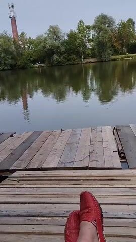 The video shows a first-person perspective of someone sitting on a wooden pier next to a calm body of water. The pier is made of weathered wooden planks. Across the water, there is a lush green shoreline with trees and a tall, red and white brick tower in the background. The sky is overcast. The person is wearing red loafers. The video is static with no discernible action.