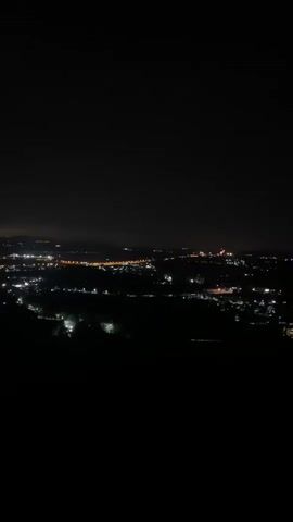 The video shows a nighttime cityscape, likely shot from an elevated position. The city lights are visible as scattered bright points across the dark expanse. The sky is black with no discernible features. Silhouetted figures of people can be seen in the foreground, suggesting the viewer is on a hill or high point looking over the city. Music is playing in the background.