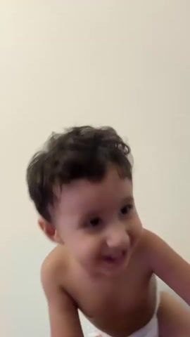 The video features a close-up of a young child, likely a toddler, with dark, curly hair. The child is shirtless and appears to be wearing white underwear. The background is a plain, light-colored wall, suggesting an indoor setting. The child is looking directly at the camera and has a wide, joyful smile, showing their teeth. Their eyes are bright and expressive, conveying happiness and excitement. The child's head is tilted slightly, and their body is angled, giving the impression they are leaning in or interacting with the camera. The lighting appears to be natural and soft, casting gentle shadows on the child's skin. The overall mood is cheerful and playful, capturing a moment of pure childhood delight. There is no visible text in the video. The time of day is not discernible, nor is there any indication of weather conditions. Given the provided location context of Sabunchu Raion, Azerbaijan, the scene is likely within a home or a similar domestic environment in that region.