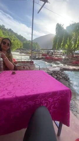 The video shows a person sitting at a table covered with a bright pink tablecloth, looking out at a view of a rushing river. A woman wearing sunglasses is seated at the table. Beyond the table, the river flows rapidly over rocks and small platforms with red chairs and tables set up for dining. Cars are parked in the background, and green trees cover the surrounding hills. The sun shines brightly, creating a slightly hazy atmosphere. A hand places a bottle of water on the table. The video is taken from a low angle, emphasizing the table and the river view.