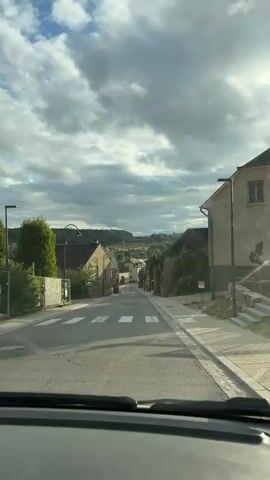 The video shows a view from inside a vehicle driving down a narrow, paved road in a village. The road is lined with buildings made of stone and stucco, some with dark tiled roofs. A pedestrian crossing is visible in the foreground. Trees and vegetation are present along the sides of the road and in the background, where rolling hills can be seen under a cloudy sky. There are no people or animals clearly visible in the footage. The buildings appear to be residential or perhaps small commercial establishments. The overall impression is of a quiet, rural European village.