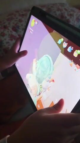 The video shows a close-up of a person's hands holding a tablet computer. The tablet screen displays a colorful video game with animated characters. One character, dressed in light blue and white, is positioned in the center of the screen, facing left. Another character, wearing a green outfit, is to the right of the first character, also facing left. The game environment appears to be indoors, with furniture and decorative items visible. The person's fingers are touching the screen, indicating they are interacting with the game. The background of the scene is blurry, but appears to be a bed with a floral patterned comforter. The lighting is dim, with the tablet screen being the primary light source.