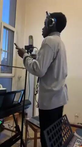 A slender, young African American man stands facing a microphone in what appears to be a modest home studio. He is wearing a gray, long-sleeved turtleneck shirt and dark pants. Headphones are perched on his head. He is holding a smartphone in his hands and appears to be reading lyrics or notes from the screen. The microphone is mounted on a stand in front of him. A keyboard, a laptop on a small wooden table, and a music stand are visible in the foreground. The room is illuminated by natural light coming in through a window on the left side of the frame. The overall impression is that the man is recording music in a simple, personal setting.