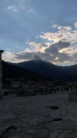 The video shows a view of a harbor town against the backdrop of a mountain range. The sky is filled with clouds, and sunlight filters through them. The town is nestled at the foot of the mountains, with boats docked in the harbor. The foreground is a stone-paved area. At the start, the camera is pointing up towards the sky and mountains but then levels to include the harbor in the shot. Toward the end, a clenched hand enters the lower left frame and exits.