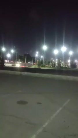 A night scene shows a large building with red lights outlining its roof. The building is illuminated by streetlights and appears to be a stadium or arena. There are cars parked in front of the building. The foreground shows a parking lot with painted lines and a manhole cover. The overall impression is of a well-lit public space at night.