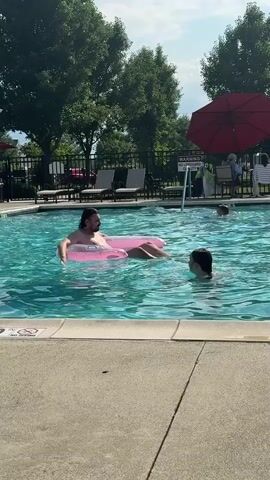 A man is floating on a pink inflatable tube in a swimming pool. Another person is in the water near him, partially submerged. The pool is surrounded by a concrete deck. In the background, there are lounge chairs, trees, a fence, and a building. The sky is blue with some clouds.