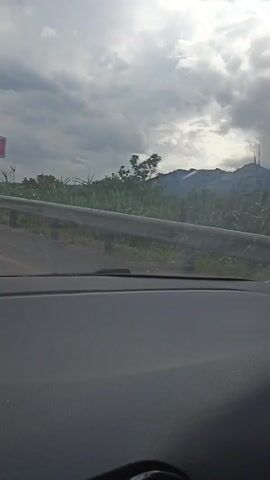 The media file captures a view from inside a vehicle, looking out through the windshield. The scene is dominated by a cloudy sky, suggesting overcast weather. A guardrail runs along the side of the road, partially obscuring the view of the landscape beyond.

The primary subjects are the natural scenery and the road. A road sign indicating a sharp left turn is visible on a pole on the left side of the frame in the initial frames. The landscape consists of rolling hills covered in dense vegetation, with some trees and possibly crops. In the background, mountain ranges are visible under the cloudy sky. The terrain appears somewhat rugged and mountainous.

The activity taking place is travel by car. The perspective from inside the vehicle indicates that the car is in motion, moving along a road that appears to be in a rural or semi-rural area. The presence of the guardrail and the road sign confirms this.

Notable details include the overcast sky, which casts a diffused light over the scene, making it difficult to determine the exact time of day, though it appears to be daytime. The location is contextually identified as Змейка, Russia. There is no visible text in the media file beyond the road sign's graphic symbol. The overall mood is subdued, likely due to the cloudy weather, and the focus is on the journey through this natural landscape.