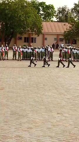 This media file captures a scene of individuals in uniform participating in what appears to be a drill or parade. The subjects are primarily people, dressed in green and white uniforms. Some are in the foreground, facing away from the camera, while others are in formation in the background, marching in unison.

The scene is set outdoors on a paved area, likely a parade ground, with buildings in the background. The architecture of the buildings suggests a warm climate, and the presence of trees indicates an outdoor environment. The time of day appears to be daytime, possibly late morning or early afternoon, given the bright lighting and shadows. The weather seems clear and fair.

The activity taking place is a synchronized march or drill. The individuals in the foreground are observing the marching group, suggesting they might be instructors or part of a reviewing party. The marching individuals are moving in a disciplined manner, their steps appearing to be coordinated. There are no explicit displays of emotion, but the overall atmosphere suggests a formal event.

The location is identified as Ajiwa, Nigeria. This context helps understand the setting, the uniforms, and the general environment depicted in the media file. There is no visible text within the media file itself. The focus is on the human activity and the organized nature of the event.