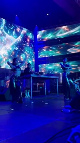 The video shows a stage performance with two dancers in sparkly outfits and a DJ behind a table. The background is a large screen displaying a cosmic or galaxy-themed visual with bright lights and colors. The dancers are performing synchronized movements. The stage is lit with blue and purple lights.