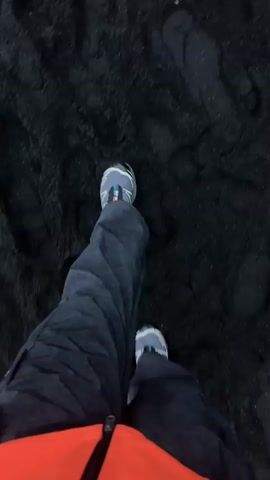 Here's a description of the video:
The video starts with a close-up view of someone walking on a black sand beach. The person is wearing dark pants, a bright orange jacket, and grey and white shoes. The ground is covered in coarse black sand, and there are visible footprints. As the video continues, the camera pans up to reveal the broader scene. The black sand beach stretches out towards the ocean. In the distance, there are distinctive rock formations jutting out of the water, and a green, grassy cliff is visible along the coastline. The sky is overcast and grey.