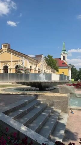 The video captures a bright, sunny day in Františkovy Lázně, Czechia. The scene is dominated by a serene park-like setting with a prominent fountain in the foreground. The fountain's water sprays upwards, creating a refreshing ambiance. Behind the fountain, a pathway leads to a long, pale yellow building with arched windows, suggesting a historical or public structure. Further in the background, a red-roofed building with a green-spired clock tower is visible, hinting at a church or a similar landmark.

The park features carefully arranged steps leading down towards the fountain, constructed from stone. Vibrant pink flowers bloom in abundance in planters and along the edges of the pathway, adding a splash of color to the scene. Hanging baskets filled with white and pink flowers are suspended from decorative poles, further enhancing the aesthetic appeal of the park. Green trees provide shade, and a clear blue sky with scattered white clouds suggests pleasant weather.

The overall impression is one of tranquility and natural beauty, ideal for relaxation and strolling. There is no visible text within the media file. The time of day is clearly daytime, indicated by the strong sunlight and shadows. The location cues, such as the architectural style of the buildings and the park's layout, are consistent with a European spa town. The lack of people suggests a peaceful moment captured, perhaps early in the morning or during a quiet part of the day. The scene evokes a sense of calm and well-being.