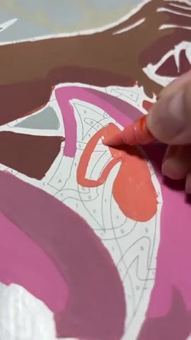 A close-up shot shows a hand holding a pink marker and coloring in a section of a paint-by-numbers canvas. The canvas is partially filled with colors, including pink, brown, and a lighter shade of pink. Numbers are visible on the canvas, indicating where to paint. The marker is actively being used to fill in a numbered section with pink paint.