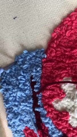 This is a close-up video of someone punch-hooking a piece of fabric with yarn. The fabric is a textured canvas, and the yarn is thick and plush. The design being created appears to be a colorful abstract pattern with sections of bright pink, light blue, and white yarn. A metal punch needle tool is visible, being used to create loops of yarn on the canvas. The activity is detailed and focused, suggesting a craft project in progress. The lighting is bright, indicating daytime. There are no visible people or explicit location cues within the frame itself, apart from the implied setting of a crafting workspace. No text is visible.