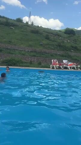 The video shows an outdoor swimming pool with a blue surface under a bright sky with white clouds. In the foreground, several people are in the water. On the left side, two people are partially visible in the pool, their heads above the surface. Further back, to the right, three people are walking by the poolside. Behind them, two individuals are relaxing on lounge chairs. The pool area is surrounded by lush green grass and vegetation, with a gentle slope rising in the background. The overall scene depicts a leisurely day at the pool.