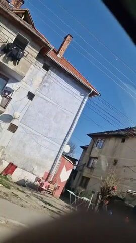 The video shows a view from inside a car, looking out onto a street scene in a residential area. Initially, a group of people, including one on a bicycle, are visible. The camera then pans upwards, showing various buildings and a utility pole against a clear blue sky. The buildings appear to be multi-story residential structures with visible details like balconies, windows, and chimneys. The utility pole is laden with wires. The camera movement is continuous, providing a moving perspective of the surroundings.