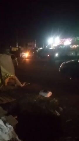 The video, taken at night, shows a road with several vehicles, including cars and a van, parked on the road. The area is illuminated by streetlights, which are bright, and some festive lighting is also visible. There's a dark foreground, with some scattered debris and overgrown vegetation. A makeshift shelter or covering is visible to the left. The overall scene is slightly blurry.