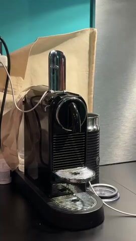The camera focuses on a black Nespresso coffee machine placed on a black surface. To the right of the coffee machine is a black milk frother. In the background, there is a beige canvas and a metal-like surface. A white cable snakes across the surface to the left of the coffee machine, and a small, white container is also visible. The background includes a blue-green painted wall. The lighting is adequate, providing clear visibility of the objects. There is no visible activity in the scene.