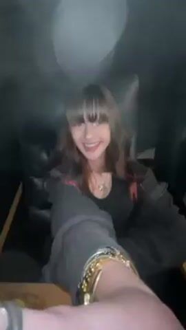 A woman with dark hair and bangs, wearing a black top and silver bracelets, is seated in what appears to be a car or limousine. She is smiling and looking at the camera, holding her arm out to show off the bracelets. The camera pans down to a tray table with yellow nachos, a large bucket of popcorn with artwork on the side, and a red cup. The environment is dark, suggesting it is nighttime or a dimly lit vehicle.