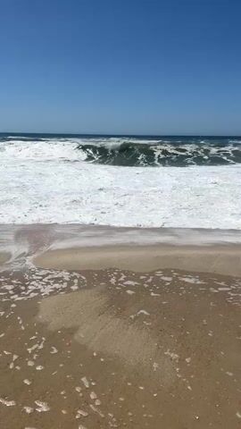 The video shows a wide shot of a beach with a clear blue sky and the ocean. Waves are crashing on the shore, creating a white foam that recedes on the sand. The sand in the foreground is wet and has some foam on it. The waves are relatively large and appear to be breaking. The sun is shining, and the overall impression is a bright, sunny day at the beach. There are no people or man-made objects visible in the frame.
