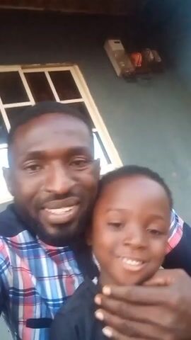 A man with a beard and a plaid shirt is holding a young boy. They are smiling at the camera. The man has his arm around the boy's neck, but does not appear to be choking him. There is a window behind them, and what appears to be an electrical box on the wall. The background appears to be a wall with some texture.