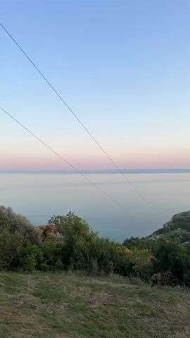 This is a vertical, static video capturing a coastal landscape at dusk. The sky transitions from a pale blue at the top to soft pinks and oranges near the horizon. Below the sky is a vast, calm body of water, likely the sea, stretching to the horizon. In the foreground, a grassy slope slopes downwards, covered in patches of dry grass and some green vegetation. Above the slope, there is a dense area of green trees and bushes. Two thin, parallel wires stretch diagonally across the upper part of the frame, originating from the left and extending towards the right, disappearing out of frame. The overall impression is one of tranquility and natural beauty as day transitions into night.