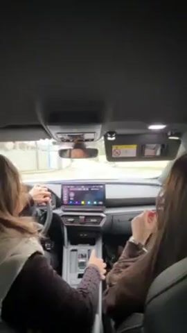 The video shows two women in the front seats of a car, likely the driver and a passenger. The driver is turning the steering wheel and touching the gear shift. The dashboard has a large touchscreen display. The car appears to be moving on a street. The surrounding environment is visible through the windshield.