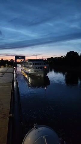The video shows a cruise ship entering a lock at dusk. The sky is a deep blue with streaks of orange and pink from the setting sun. The ship is illuminated by its own lights and the lights of the lock. The water in the lock is dark and reflects the ship and the sky. A concrete pier is visible on the left side of the screen, and trees and buildings can be seen in the background. The video appears to be shot from a stationary position on the pier.