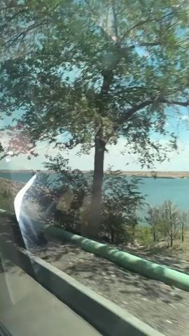 This is a video taken from inside a vehicle looking out a window. The scene shows a body of water, likely a lake or river, under a bright blue sky with some clouds. The shore is lined with sparse vegetation and rocky terrain. In the distance, a tower-like structure is visible. Power lines stretch across the sky. The camera view is slightly obstructed by reflections on the window. The overall impression is a scenic view of a natural landscape from a moving vehicle.
