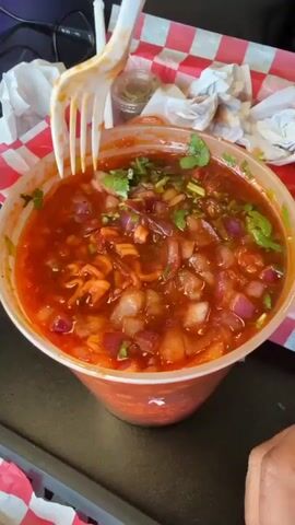 A person stirs a cup of what looks like soup with a white plastic spoon. The soup is red, with chunks of what looks like diced onions, tomato, cilantro, and noodles. The cup is plastic and slightly translucent, with the red soup coloring its lower half. It sits on a red and white checkered surface next to crumpled white napkins. A pair of glasses rests in the background to the left. The person's fingers and hand are visible as they stir the soup. The spoon moves through the soup, lifting ingredients up and mixing them.