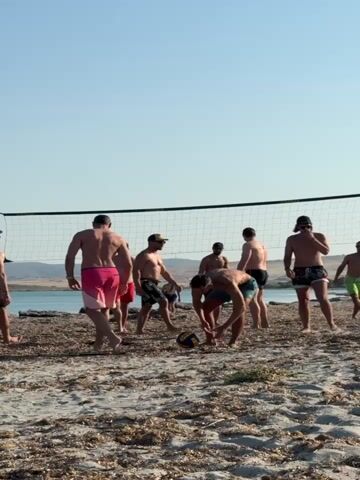 A group of men are playing beach volleyball on a sunny day. The game appears to be informal, with the players scattered across the sandy beach. The net is set up near the water's edge, and a volleyball is in play. The men are dressed in swim trunks, and most of them are shirtless, showcasing their athletic physiques. The atmosphere seems relaxed and joyful, with the players engaged in the sport and enjoying each other's company. The background shows a body of water, possibly the sea, and a hilly landscape under a clear blue sky. The time of day appears to be late afternoon or early evening, given the warm lighting and the long shadows. The presence of sand, water, and clear skies suggests a coastal or beach setting in Greece, as indicated by the location context. No visible text is present in the image.
