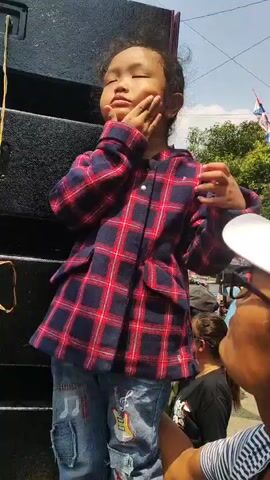 A young girl stands on top of a large black speaker, wearing a red and blue plaid jacket and blue jeans. Her right hand is raised to her forehead, and she appears to be shielding her eyes or perhaps feeling unwell. Her left hand is also raised, with her index and middle fingers extended in a V-shape. She has dark, curly hair and her eyes are partially closed. Behind her, other people are visible in the background, and there are trees and what appears to be a bright sky. The overall environment suggests an outdoor event or gathering.