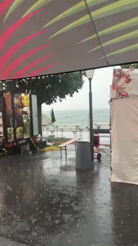 The video shows a street scene with several food stalls and a food truck. It appears to be raining heavily, with puddles of water visible on the ground and rain streaks in the air. The stalls include a crepe stand and a Thai food stand. The Thai food stand is labeled "Suweena Fine Thai Food." In the background, there is a body of water, likely a lake or sea, and trees. The overall atmosphere is dim and wet due to the rain.