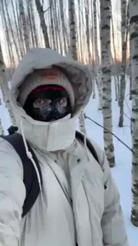 A person wearing a white winter coat with a hood, a red-and-white knit hat, and a black face mask with frozen snow on it, stands in a snowy birch forest next to a snowmobile. The snowmobile is white, red, and black, and the person is facing the camera, seemingly taking a selfie. The sun is setting in the background, casting a warm glow on the birch trees.