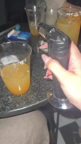 A person's hand, with long, light-colored fingernails, holds a black culinary torch with a silver tip. The torch is aimed at a tall glass filled with an orange liquid, ice, and a salt-rimmed edge. A flame emanates from the torch, igniting the salt on the rim of the glass, causing it to melt and caramelize. Two other similar glasses, also containing orange liquid and ice, are visible on the dark, speckled surface of a table. In the background, other people are present, with glimpses of their clothing and a portion of a white lace garment. A pack of cigarettes and an ashtray with cigarette butts are also on the table. The scene appears to be indoors, with a dark ambiance suggesting a bar or dimly lit gathering.