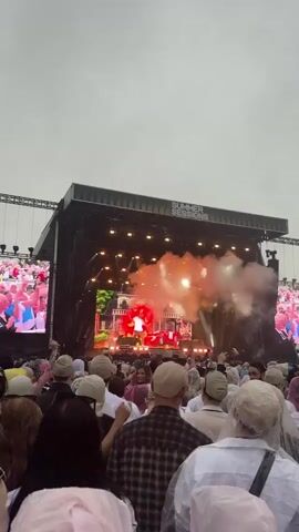 A wide-angle, vertical, POV shot captures a music concert with a large stage and a dense crowd. The stage is illuminated by bright lights and features a large screen displaying a performer. Smoke effects are visible on stage, creating a dramatic atmosphere. The crowd is facing the stage, with many people wearing light-colored hats or hoods, possibly to protect against rain. The sky is overcast and appears to be a dull gray. The video is short, showing only a few seconds of the concert.