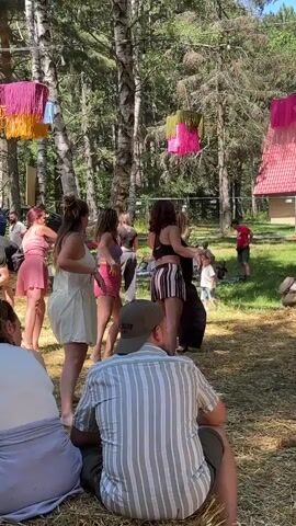 A group of women are dancing in a forest clearing. They are all facing away from the camera and are moving in sync. In the foreground, a man is sitting on a bale of hay, watching the dancers. There are trees in the background, and colorful decorations are hanging from the trees. The scene appears to be a festival or outdoor event.