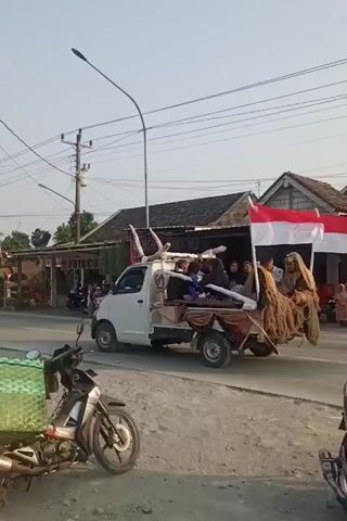 A white pickup truck is driving down a street with two Indonesian flags flying from the back. The truck is carrying several people, some of whom are wearing traditional costumes. The truck also appears to be carrying some straw or hay. There are other vehicles on the road, including motorcycles. The scene appears to be in a rural or suburban area, with shops and houses visible along the street. The sky is clear and the sun appears to be setting.
