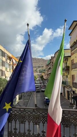 The video captures a daytime view from a balcony overlooking a street in Maddaloni, Italy. In the foreground, two flags are prominently displayed on poles, gently swaying in a light breeze. On the left is the blue flag of the European Union, adorned with a circle of yellow stars. On the right is the Italian flag, featuring vertical stripes of green, white, and red. A decorative black metal railing with an intricate pattern frames the bottom of the shot.

Below the balcony, a street stretches into the distance, with cars moving and parked along its length. Multi-story buildings, characteristic of Italian architecture with balconies and shops, line both sides of the street. In the background, a large, rocky hill or mountain rises under a bright blue sky dotted with white clouds. The scene suggests a sunny and pleasant day in an Italian town. No legible text is visible in the video.