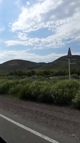 The video shows a view from a moving vehicle, likely a car, looking out the passenger side window. The road is visible in the foreground, with a gravel shoulder and a white line marking the edge of the paved surface. Beyond the road is a green, grassy field with various plants and bushes. In the middle ground, there are rolling hills covered in vegetation. The sky is partly cloudy with a mix of blue sky and white clouds. The camera pans slightly to the right, revealing more of the landscape. There are some faint, light-colored markings or structures on the side of the hill in the distance, but they are not clearly discernible. A tall, thin object, possibly a signpost with red and white markings, is visible on the right side of the frame, near the middle ground. The overall impression is of a rural or natural landscape.